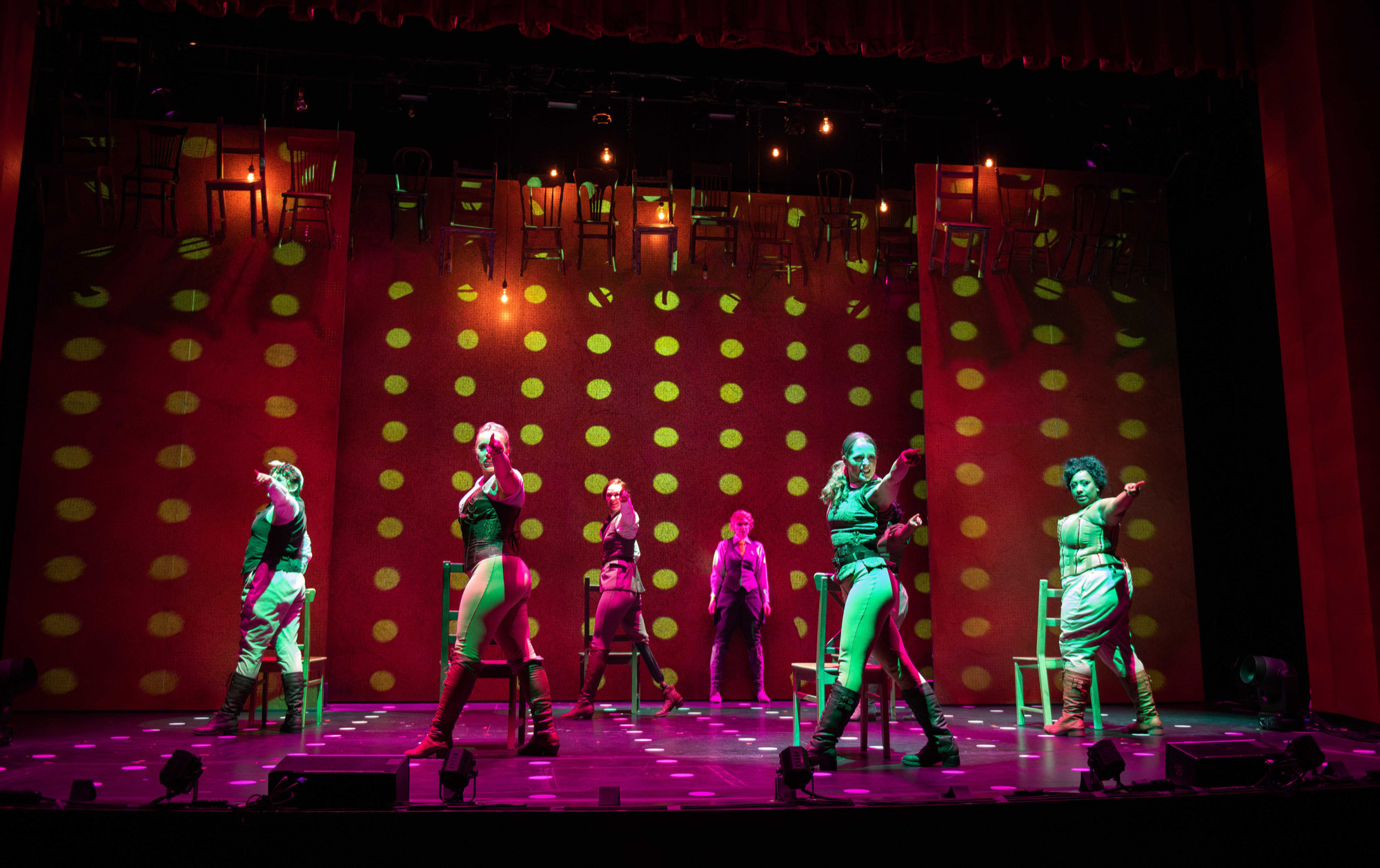 Seven women stand on stage, under moody but vibrant lights. Six stand by chairs, pointing towards the audience, while the seventh stands at the back of the stage.