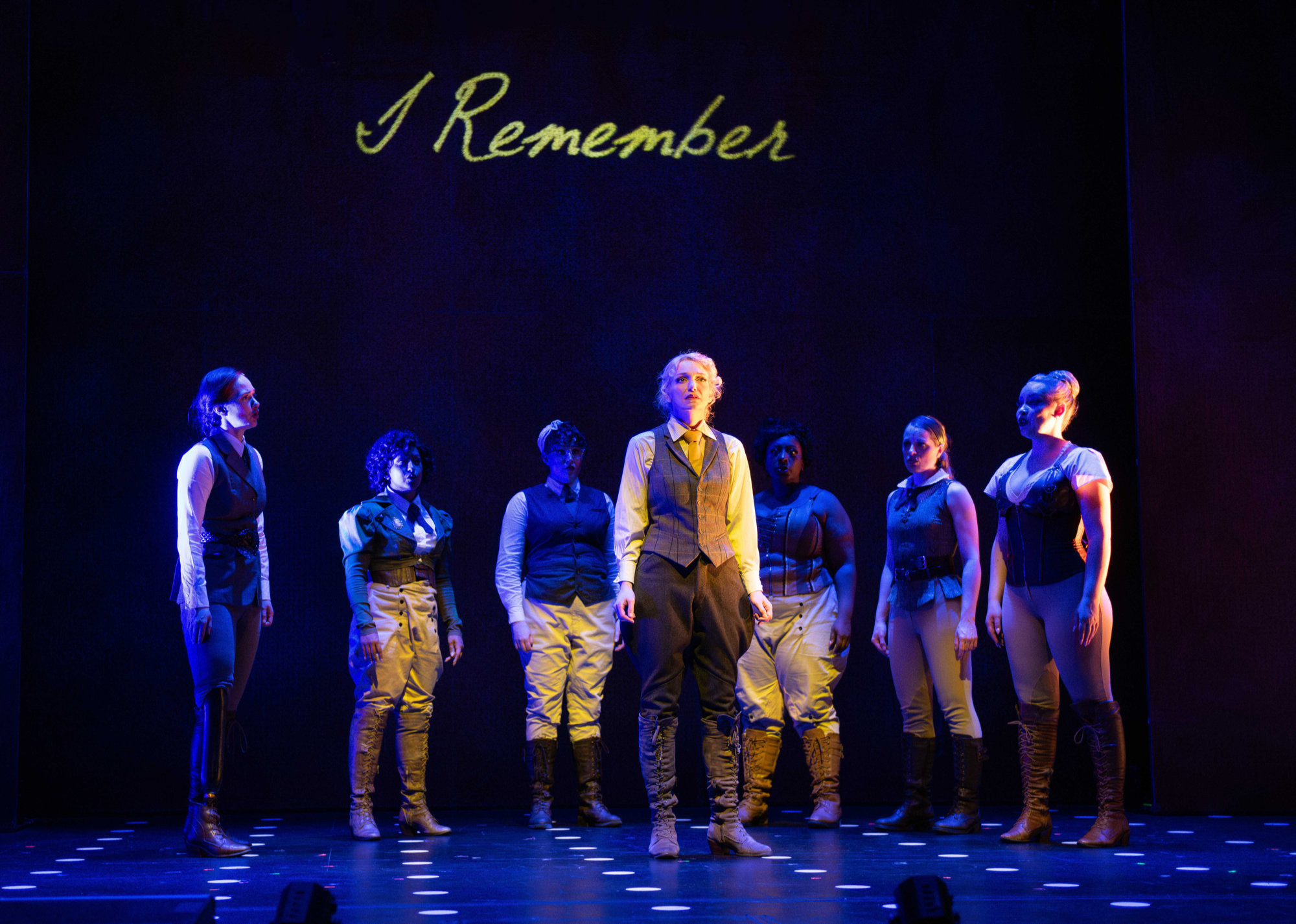 Six women stand in a semi-circle behind a seventh woman who stands in the spotlight, under a blue cast of light.