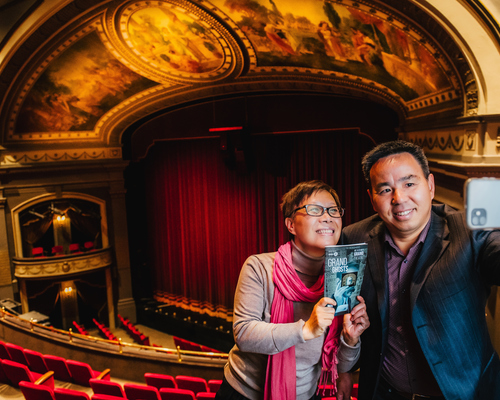 A photograph of the Spriet Stage taken from the balcony, with two individuals posing together with a house program.