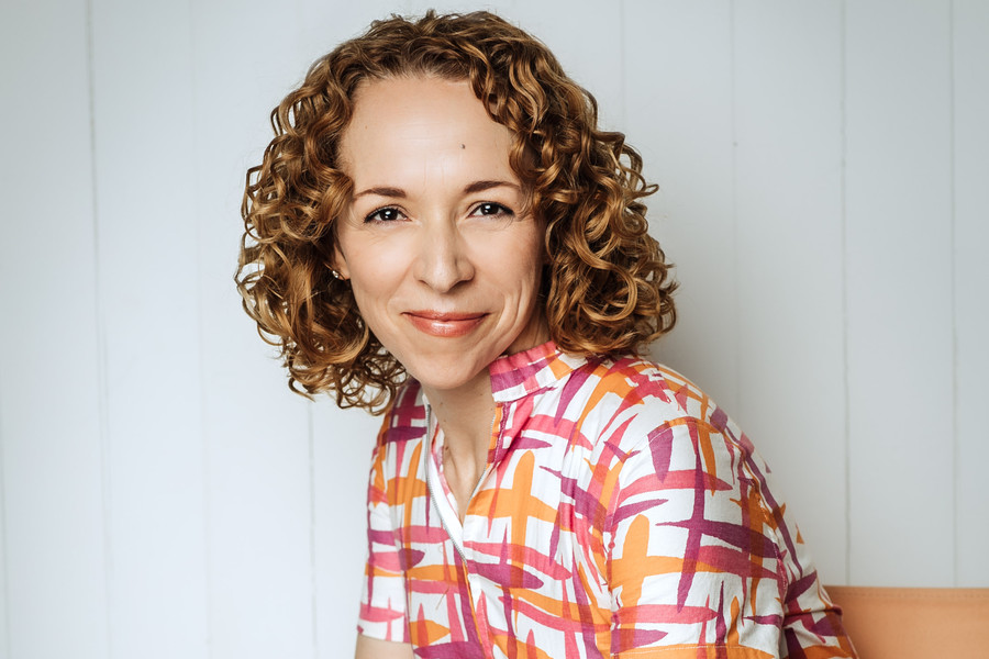 Rachel Peake sits in front of a neutral background. She wears a vibrant outfit with purple and orange details.