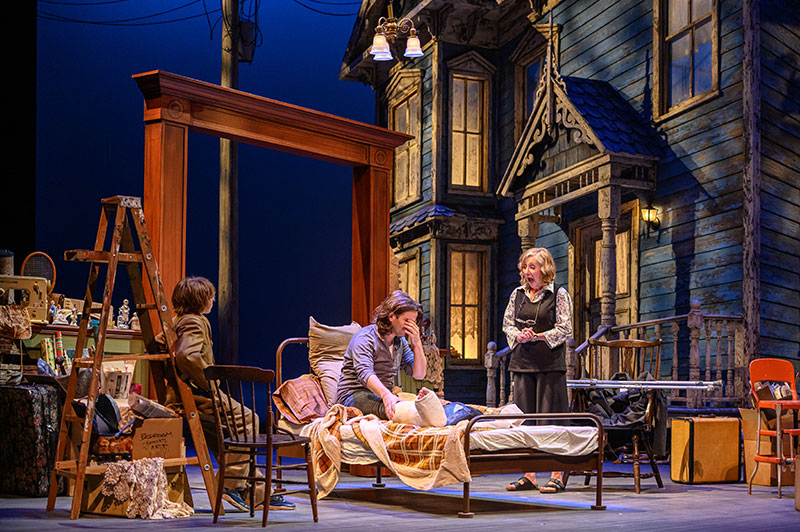 A young man with a cast on his leg sits on a bed on the set of This London Life. A young boy leans against a ladder on one side of the bed, and a woman stands on the other side, speaking.