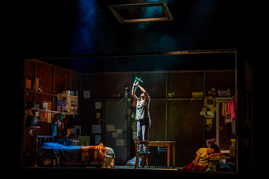 Alexis Gordon stands on a chair centre stage holding a lamp toward a skylight in the Grand Theatre's production of Room.