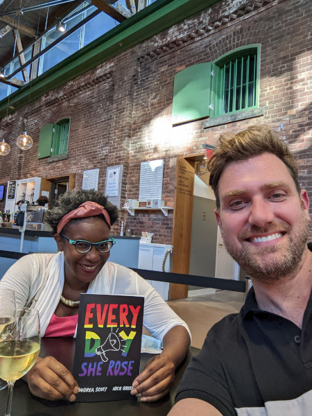 Andrea Scott and a friend smile as they sit at a table. Andrea holds a copy of her play Every Day She Rose.