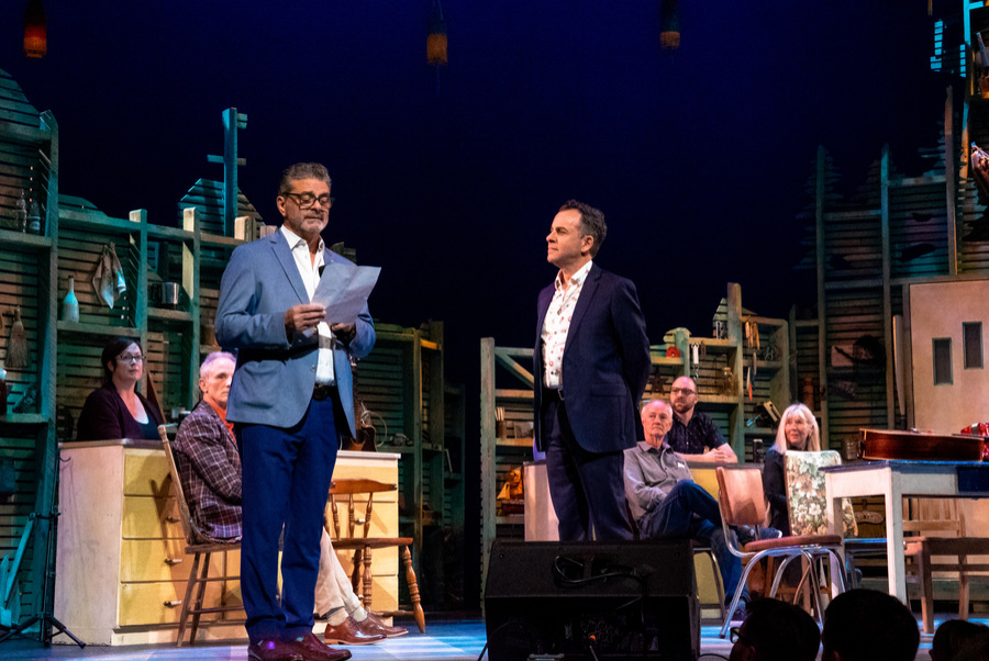 George Kerhoulas reads from a sheet of paper as he stands on the Spriet Stage on the opening night of East Coast Kitchen Party. Dennis Garnhum stands by him, and cast members are seated around the set in the background.