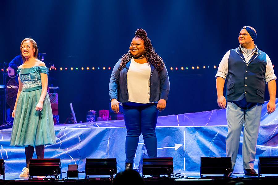 Gabi Epstein, Masini McDermott, and Jacob MacInnis smile as they stand at the footlights during a performance of Home for the Holidays.