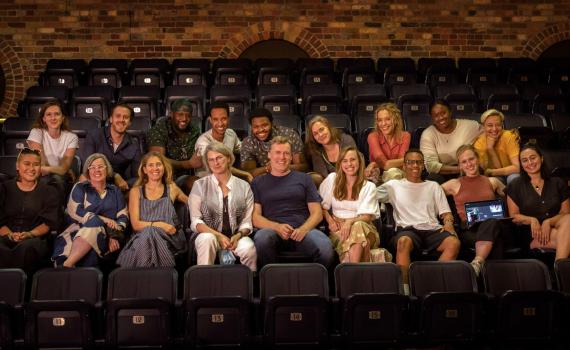 The company of Fall On Your Knees sits in two rows of an intimate theatre space and smiles at the camera. The theatre chairs are dark in colour and the back wall is a multi-shade brick. Photo by John Lauener Photography