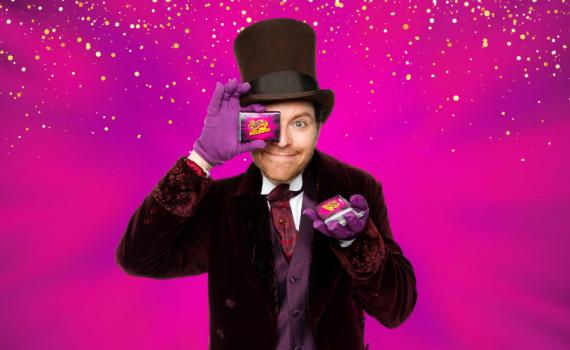 Mark Uhre as Willy Wonka holds up a chocolate bar, in front of a vibrant pink and purple background.