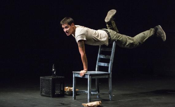 A young man balances horizontally on the back of a wooden blue chair, one hand on the seat.