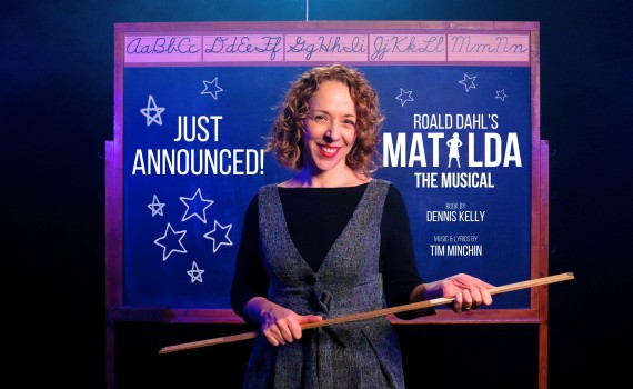 Artistic Director Rachel Peak holds a ruler as she stands in front of a chalkboard. A message on the chalkboard announces the 2024/25 season High School Project, Matilda the Musical.