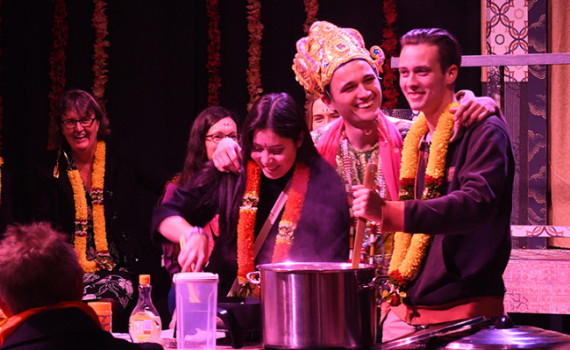 Three people wearing garlands and costumes, laughing and cooking with an audience in the background.