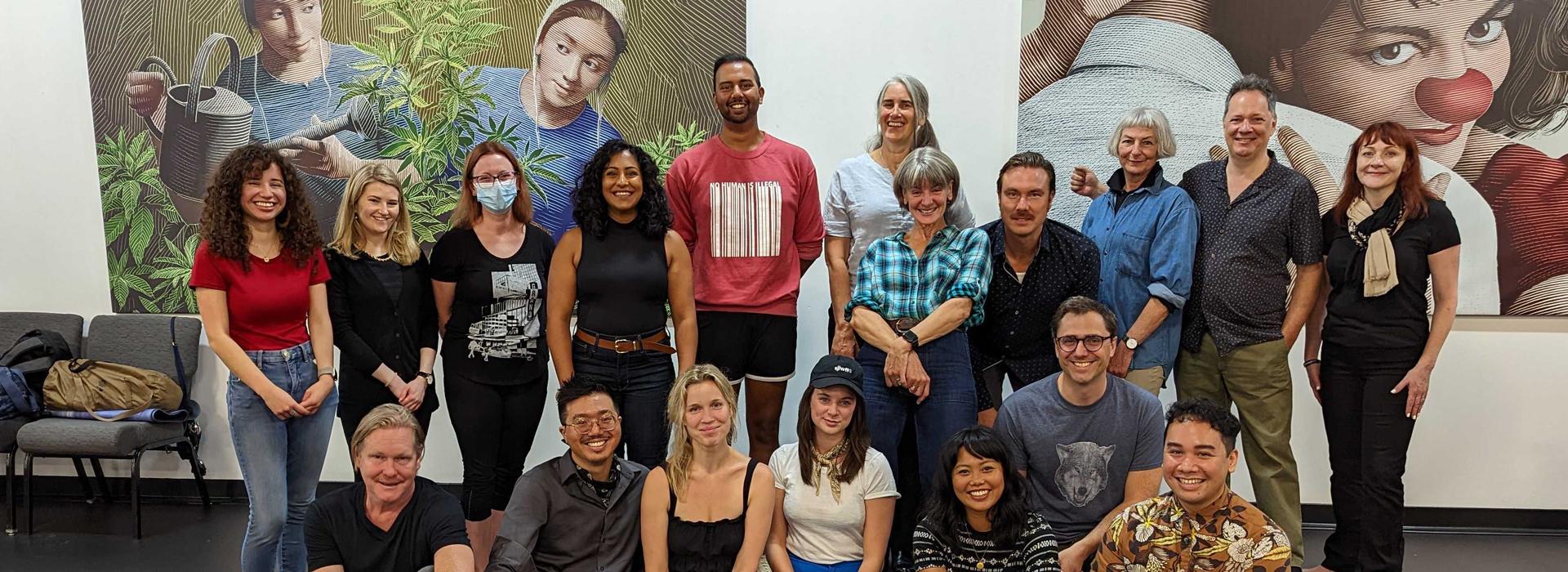 The company of Grand Ghosts (18 people) stands in a group formation in a modern rehearsal space and smiles at the camera. In the background, two large graphic posters are visible: one showcasing two Amish women watering a cannabis plant, the other presenting a woman in clown apparel hugging a man (who is only seen from behind).