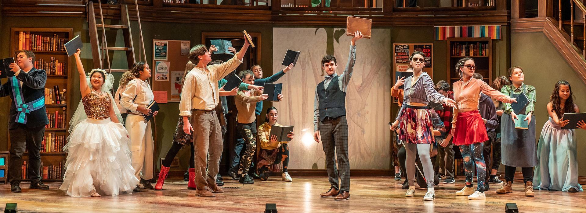 The cast of Into the Woods stands staggered on the Spriet Stage holding books triumphantly in the air. The background set emulates a library and a large plant bean stock grows in the back middle. Photo by Dahlia Katz.
