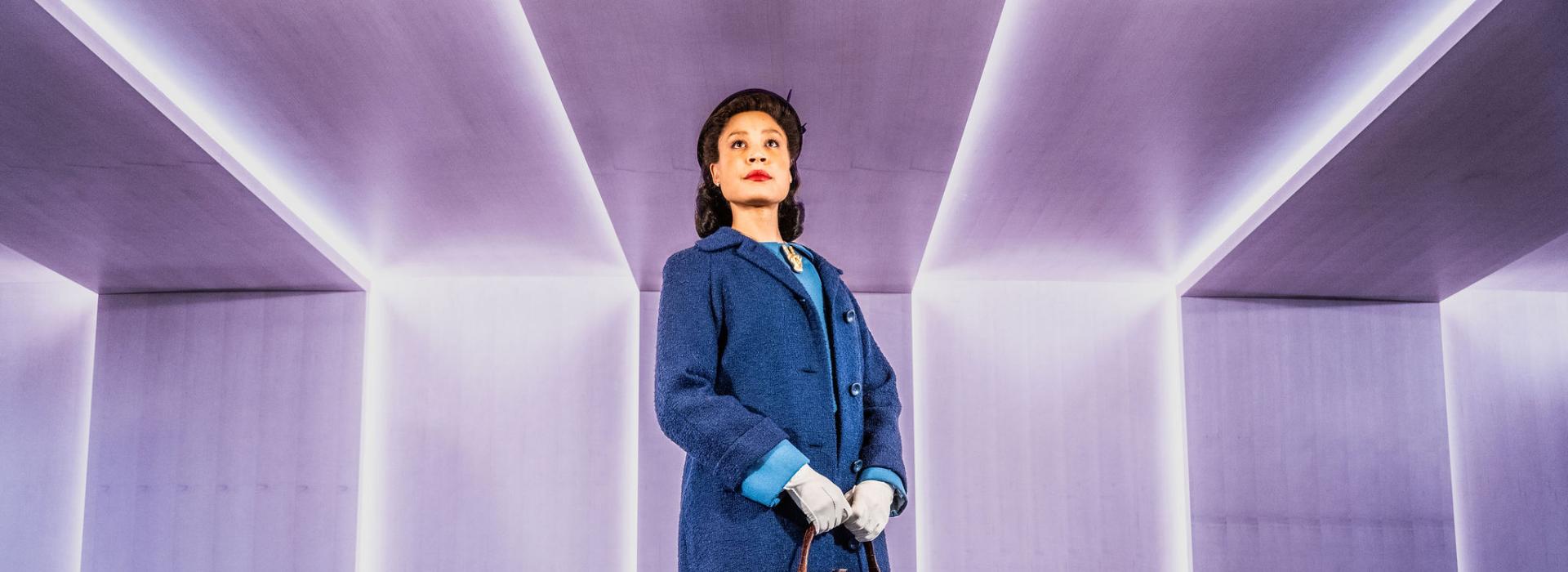 Beck Lloyd in character as Viola Desmond, stands in front of a light purple wall with vertical strips of light that continue along the ceiling.