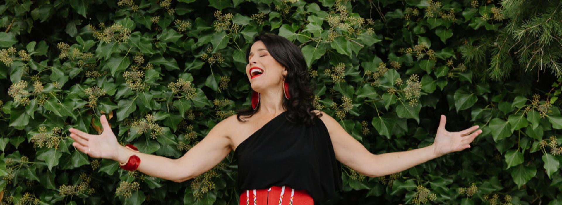 Andrea Menard stands in front of some greenery, wearing a black and red dress with her arms outstretched.
