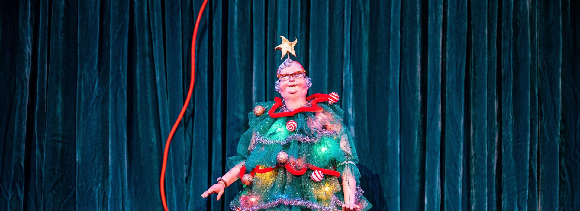 A marionette in a dress that resembles and is decorated like a christmas tree stands in front of a teal curtain.