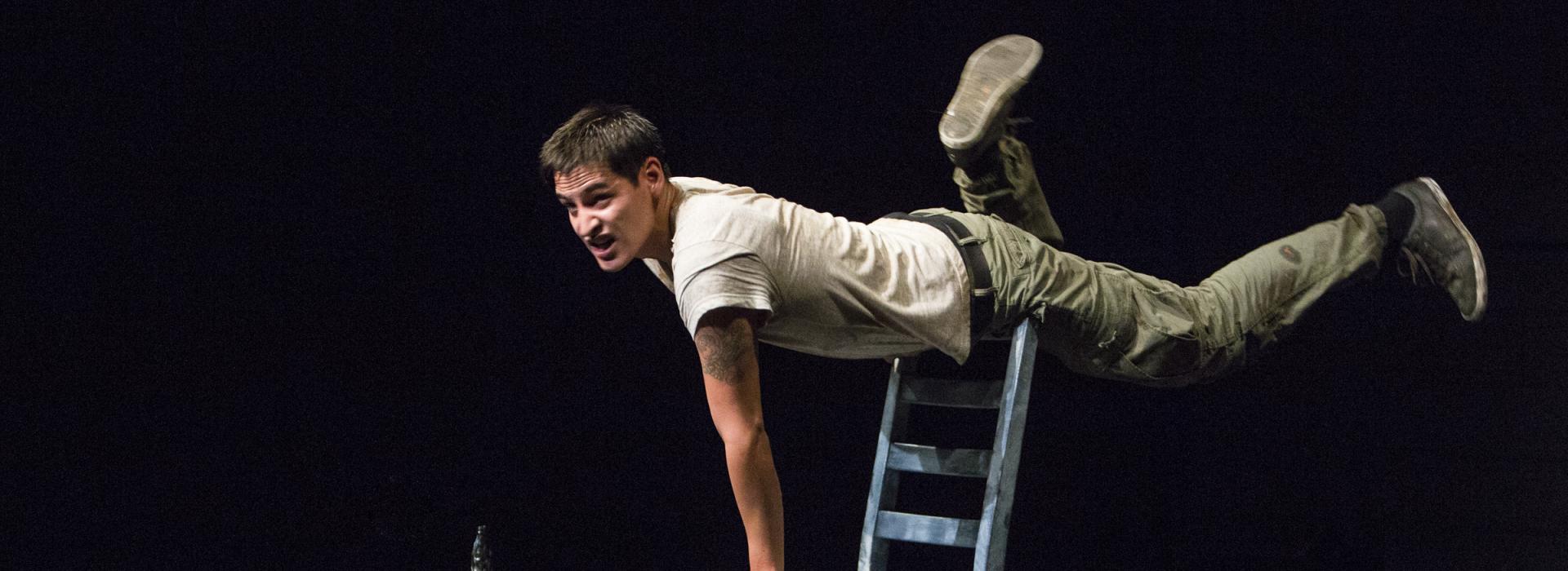 A young man balances horizontally on the back of a wooden blue chair, one hand on the seat.