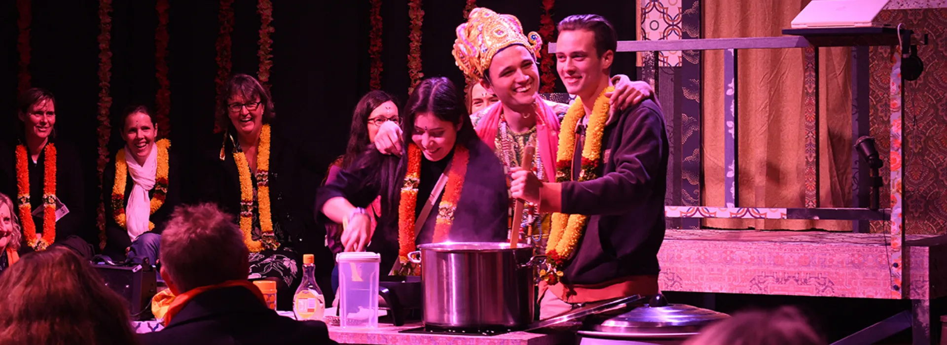 Three people wearing garlands and costumes, laughing and cooking with an audience in the background.
